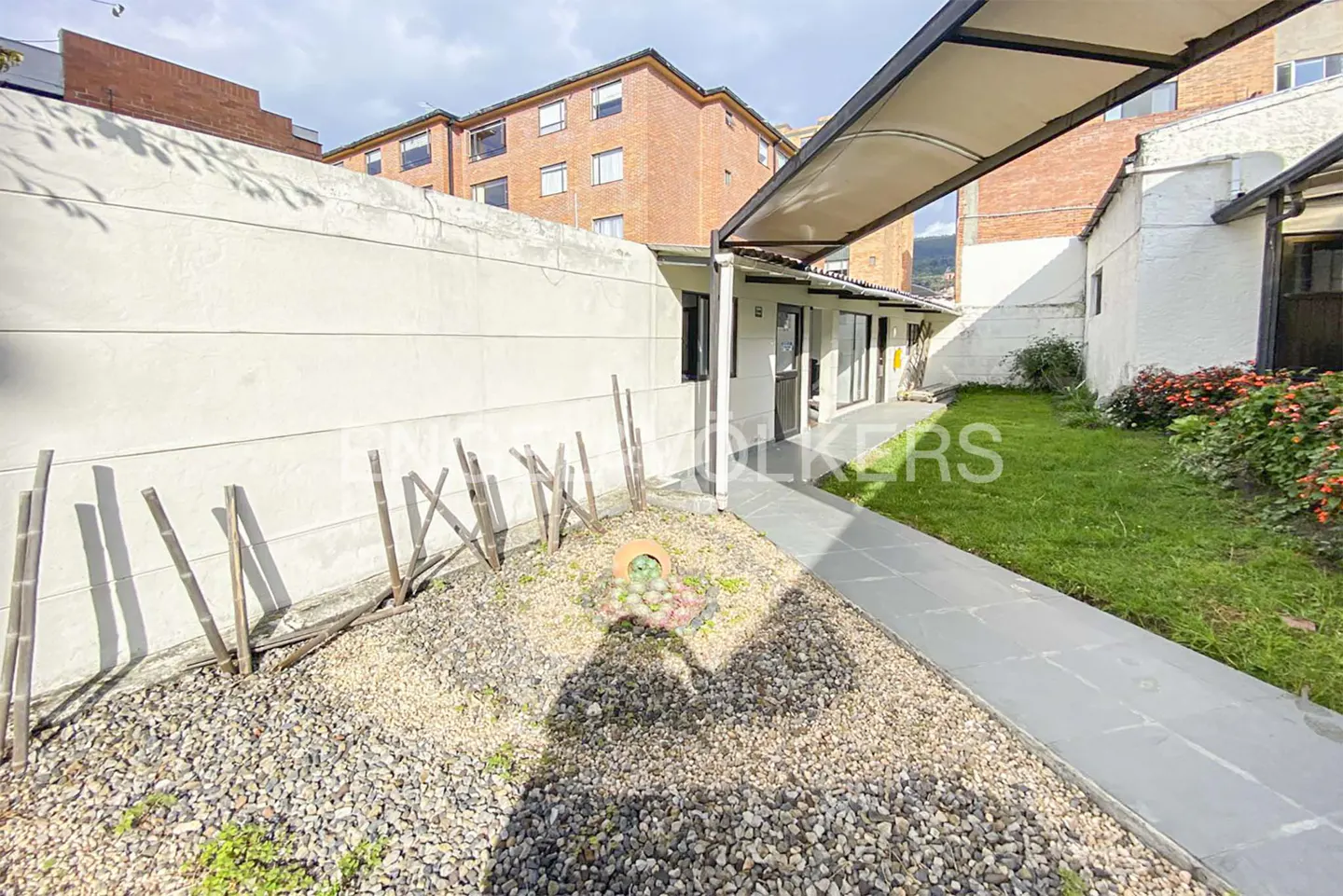 Exterior view of a building with a stone walkway, a small garden with rocks, and a white wall with bamboo sticks.