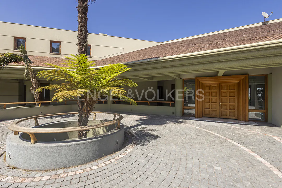 Exterior view of a beige building with a brown roof and a large wooden door. A circular planter with a tree sits in a cobblestone courtyard.