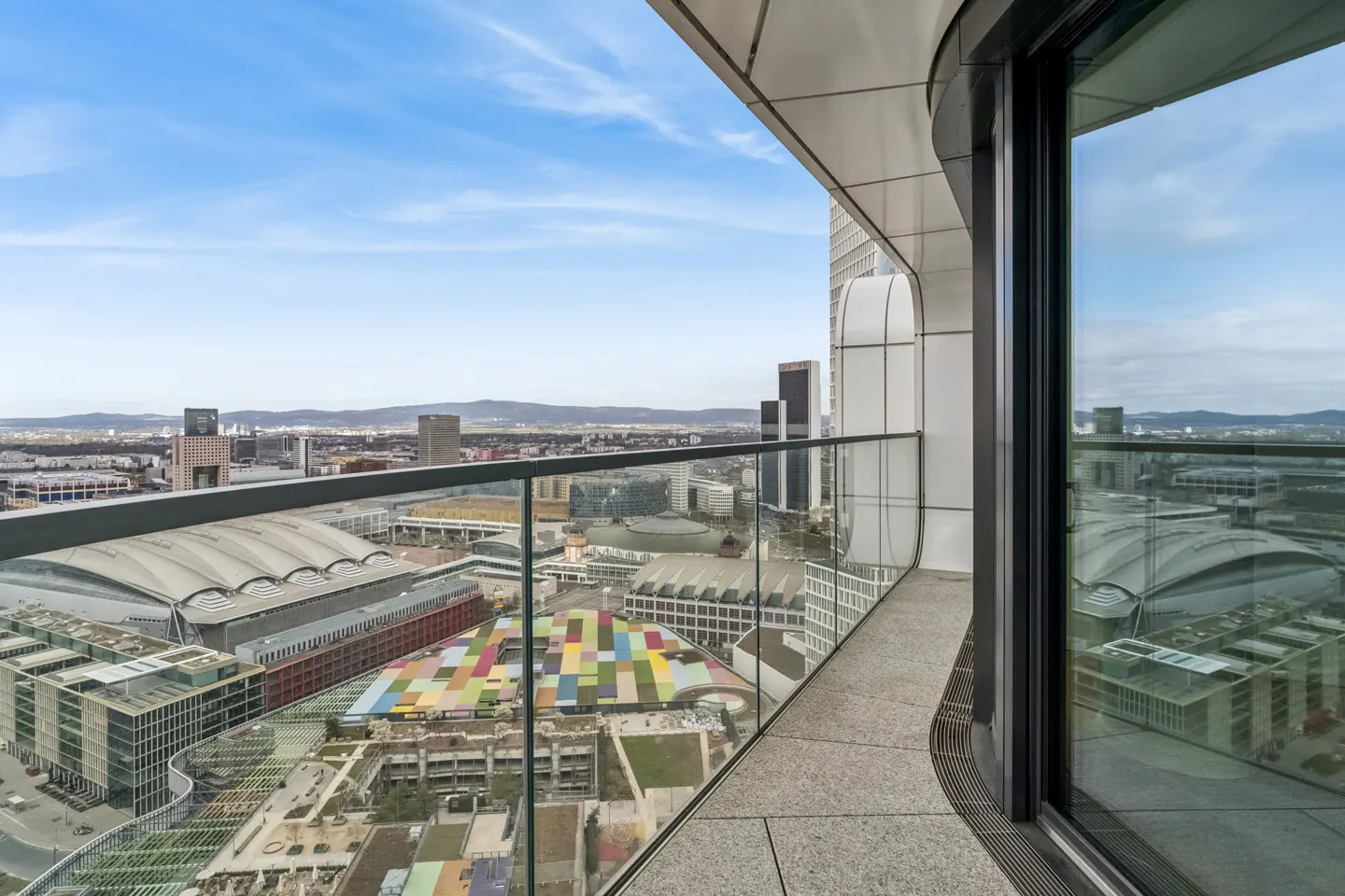 View from a high-rise balcony with glass railings overlooking a city with a colorful building roof.