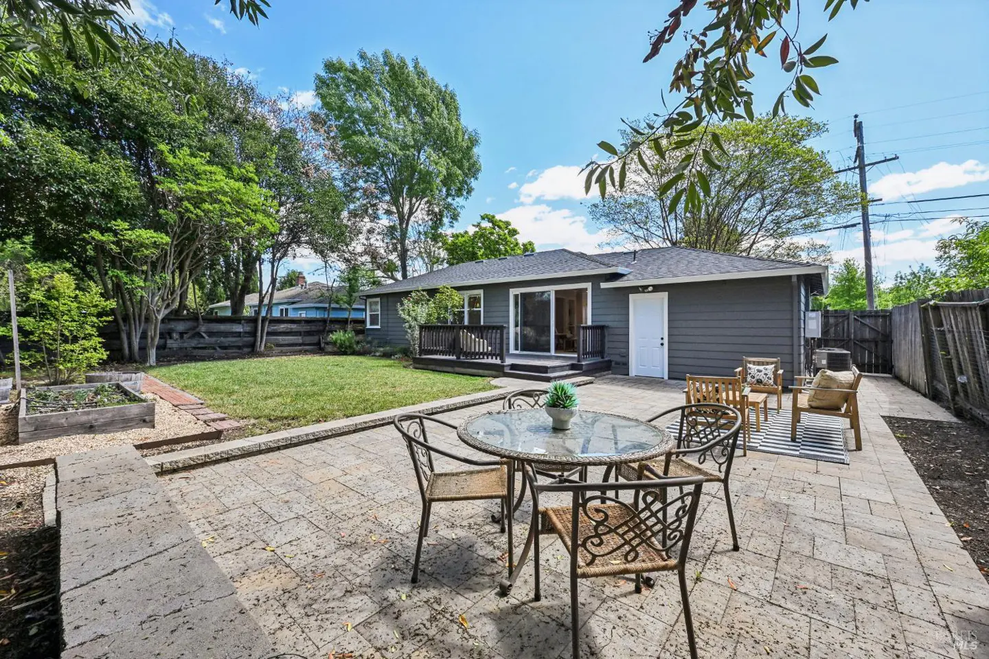 Backyard patio with a glass table and four chairs, a gray house with a deck, and green grass.