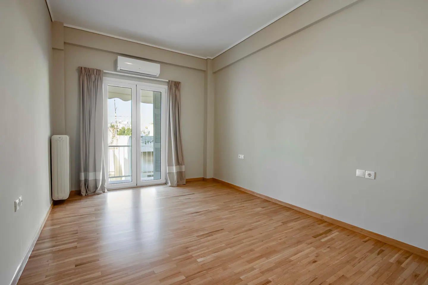 Empty room with light gray walls, wood floors, and a glass door to a balcony with gray curtains.