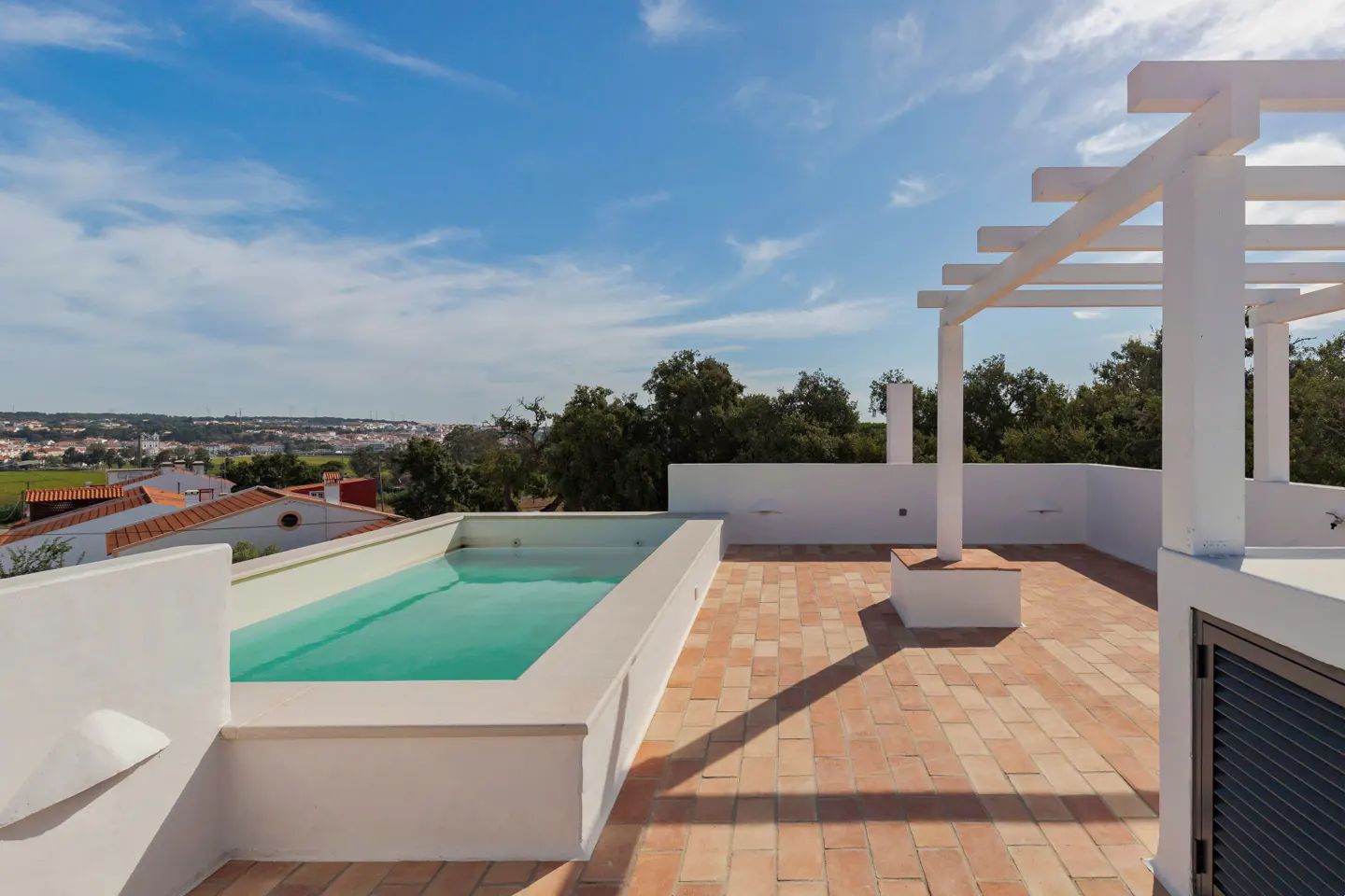 Rooftop pool with turquoise water, white walls, and brick patio. White pergola, trees, and a town visible in the background under a blue sky.