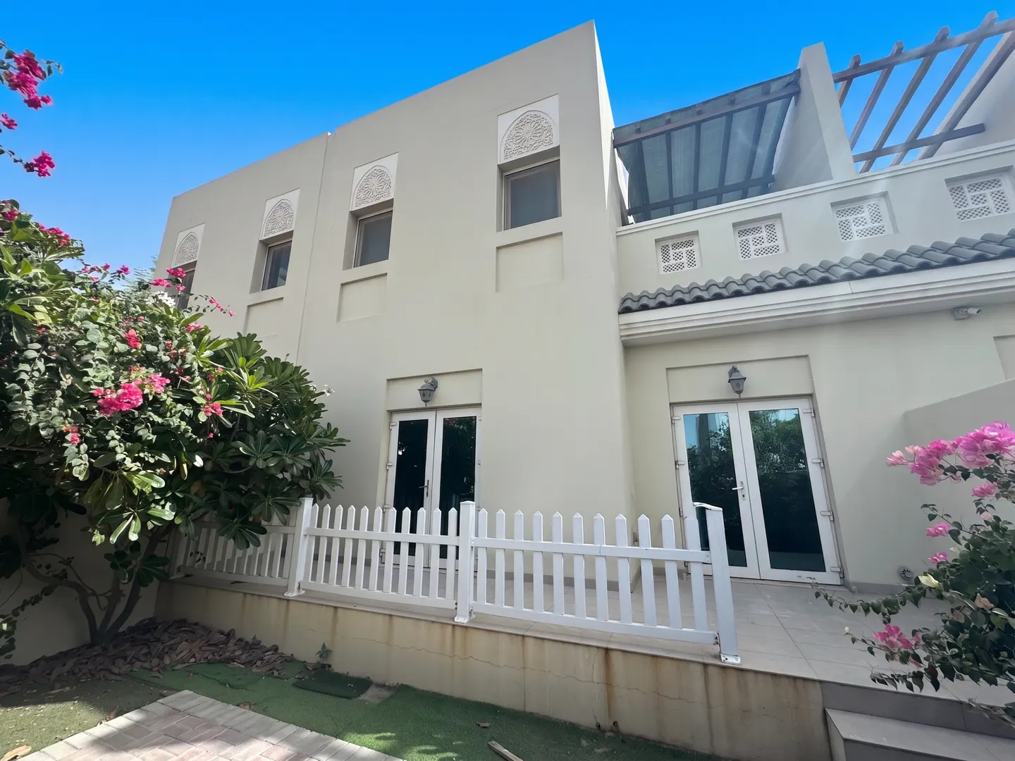 Two-story beige house with white trim, a white picket fence, and pink flowers against a blue sky.