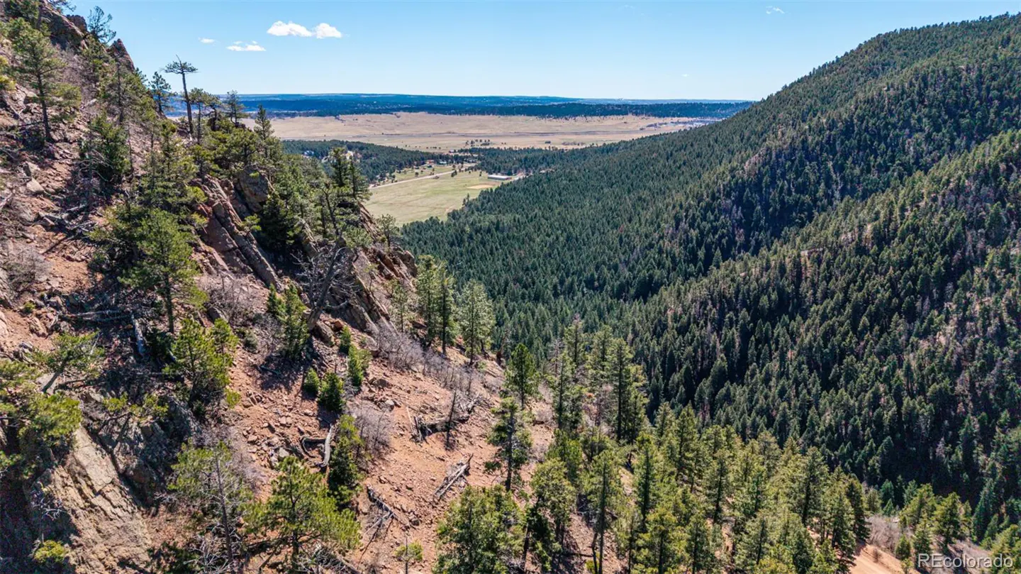 Mountain view with green trees, brown rocks, and a valley in the background under a blue sky.