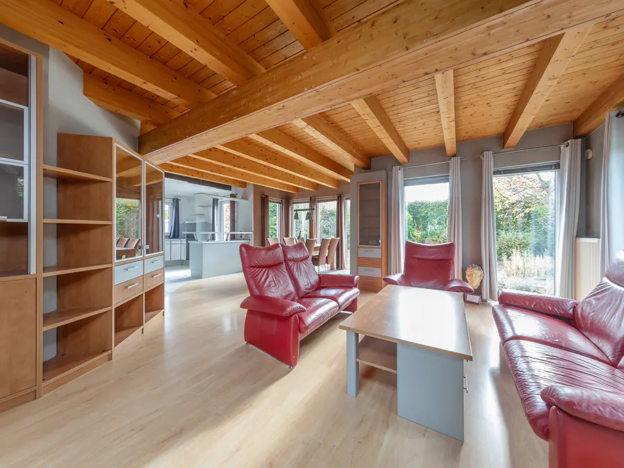 Bright living room with wood floors, exposed beams, red leather sofas, and large windows overlooking a green yard.