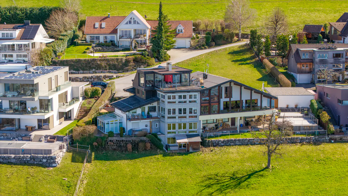 A multi-story gray house sits on a green hill, with other houses visible in the background.