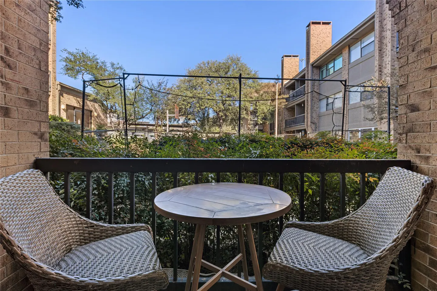 Balcony view with two wicker chairs and a small round table. Greenery and buildings are visible in the background under a blue sky.