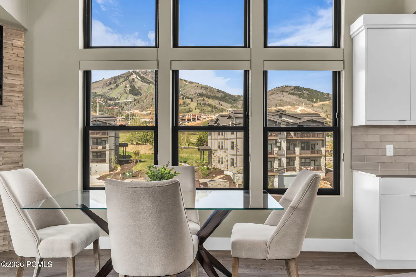 Dining area with glass table, beige chairs, and a large window view of mountains and buildings. White kitchen cabinets are visible.