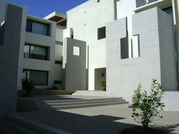 Modern white building exterior with geometric concrete accents and steps leading to an entrance. Small green plants add a touch of nature.