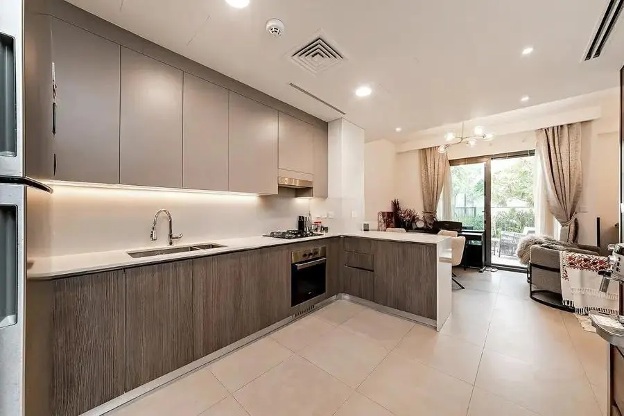 A modern kitchen with gray cabinets, white countertops, and stainless steel appliances. The kitchen opens to a living area with a sliding glass door.