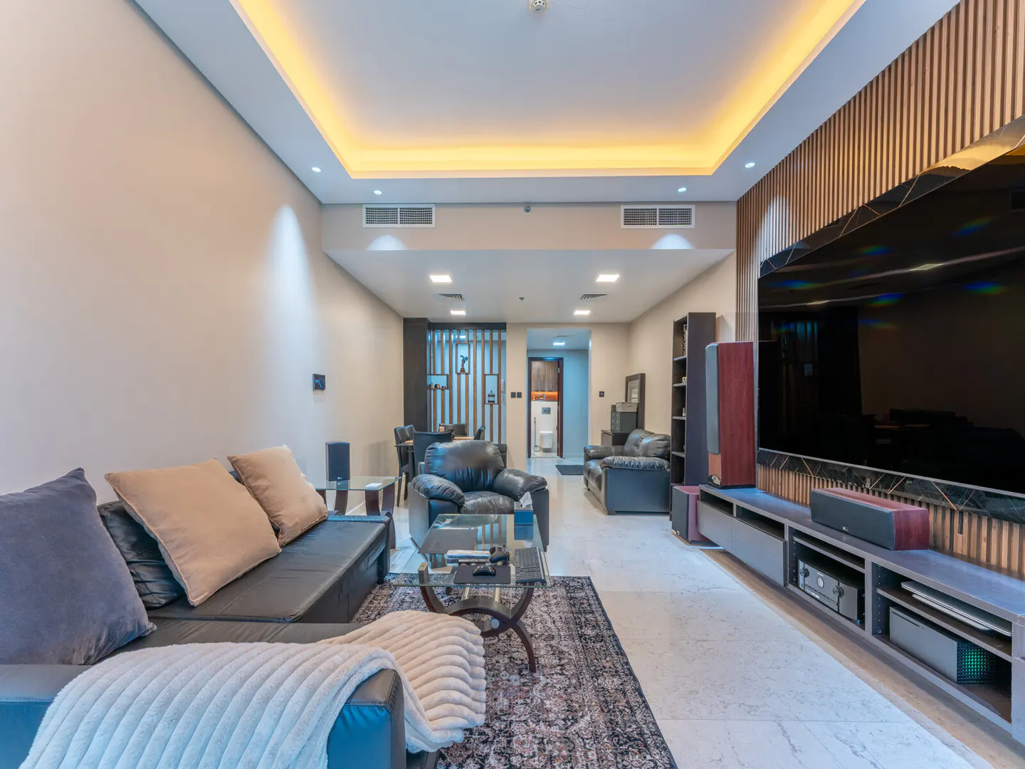 Living room with black leather sofa, armchairs, glass table, and large TV on a wood-paneled wall. Beige walls and marble floors.