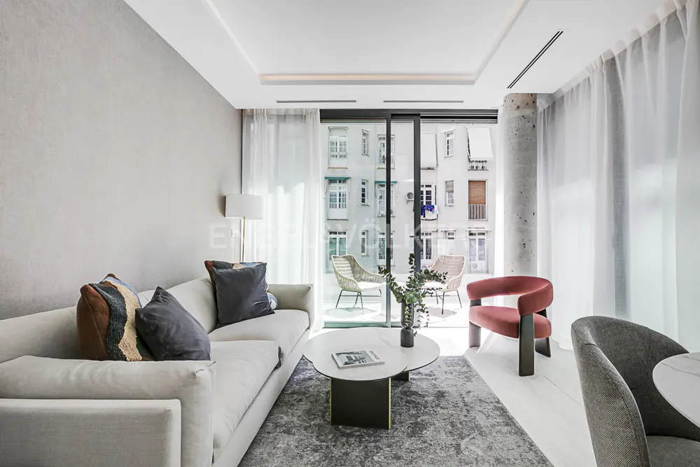 Bright living room with a white sofa, patterned pillows, and a round coffee table. Balcony with chairs visible through sliding glass doors.