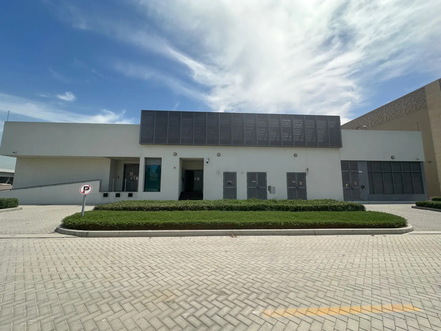 Exterior of a modern, white building with a gray facade detail, green landscaping, and a paved parking area under a blue sky.