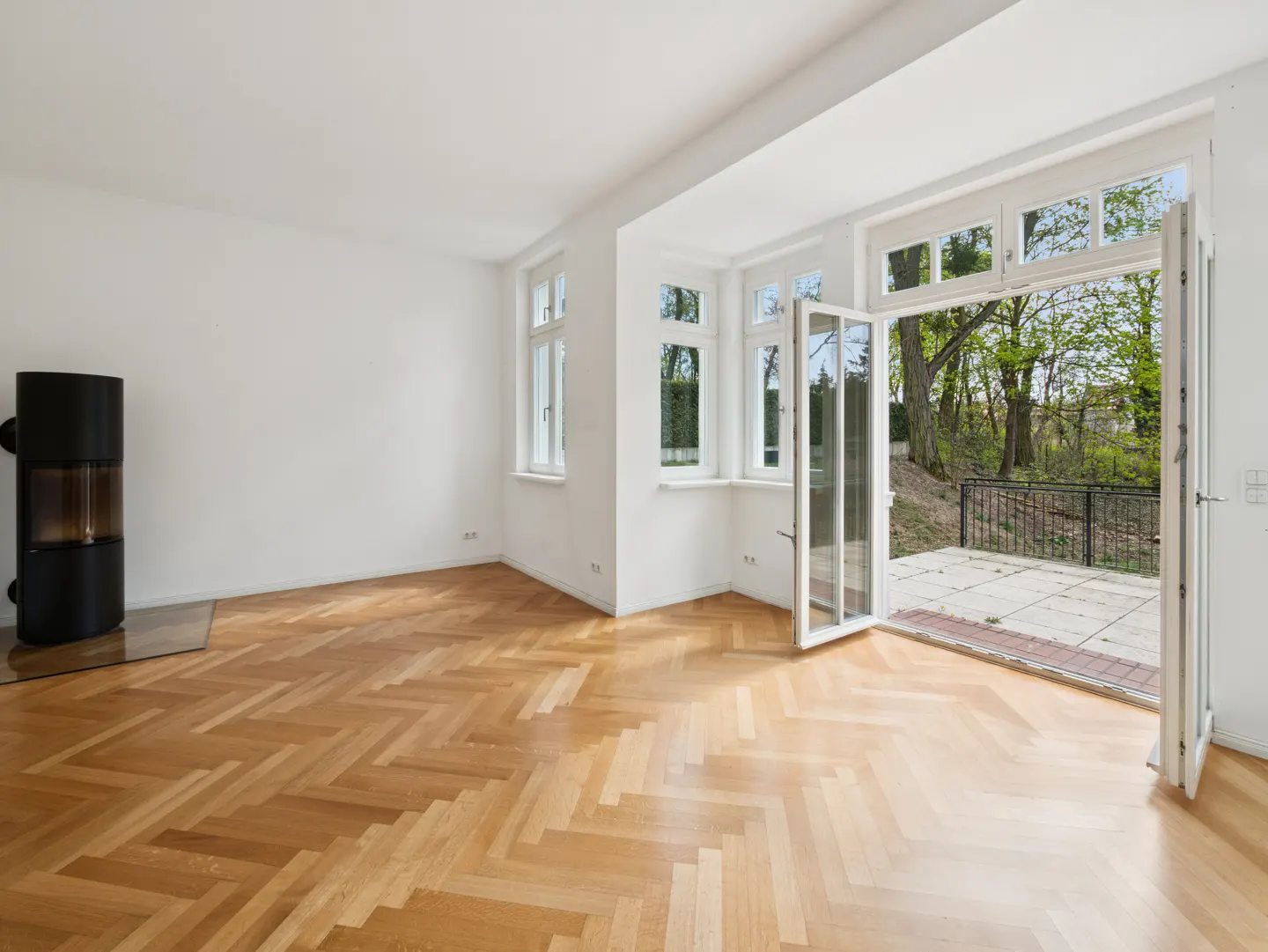 Bright, empty room with herringbone wood floors, white walls, a black stove, and open French doors to a patio.