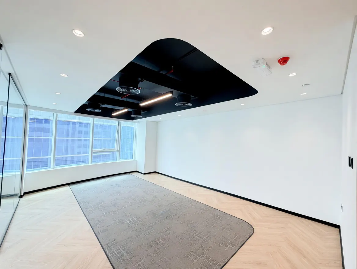 Bright, empty office space with large windows, white walls, and a gray rug on a wood floor. A black, curved ceiling feature holds lights and vents.