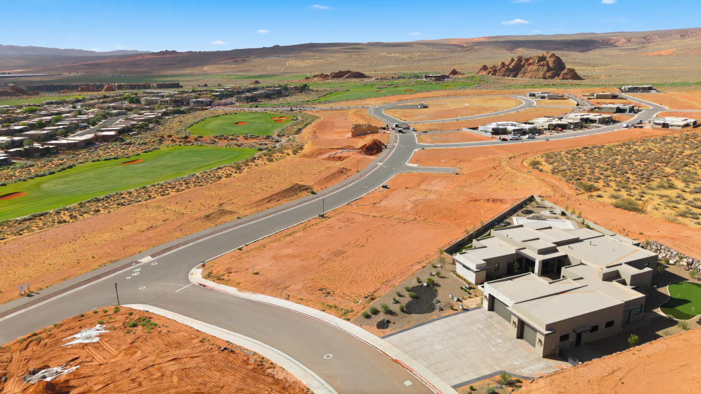 Aerial view of a modern home in a desert landscape with red soil, a golf course, and mountains in the background.