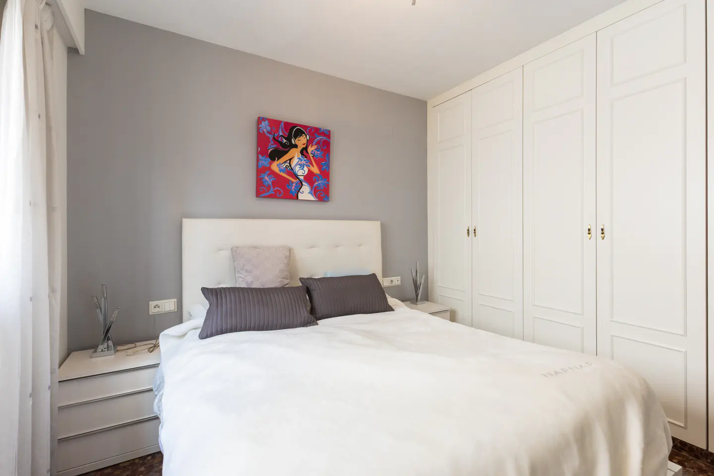 Bedroom with a white bed, gray pillows, and a colorful painting on a gray wall. White wardrobe on the right.