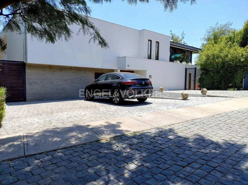 Modern white house with a black Mercedes parked in the driveway. The driveway is paved with gray stones. Trees are visible in the background.