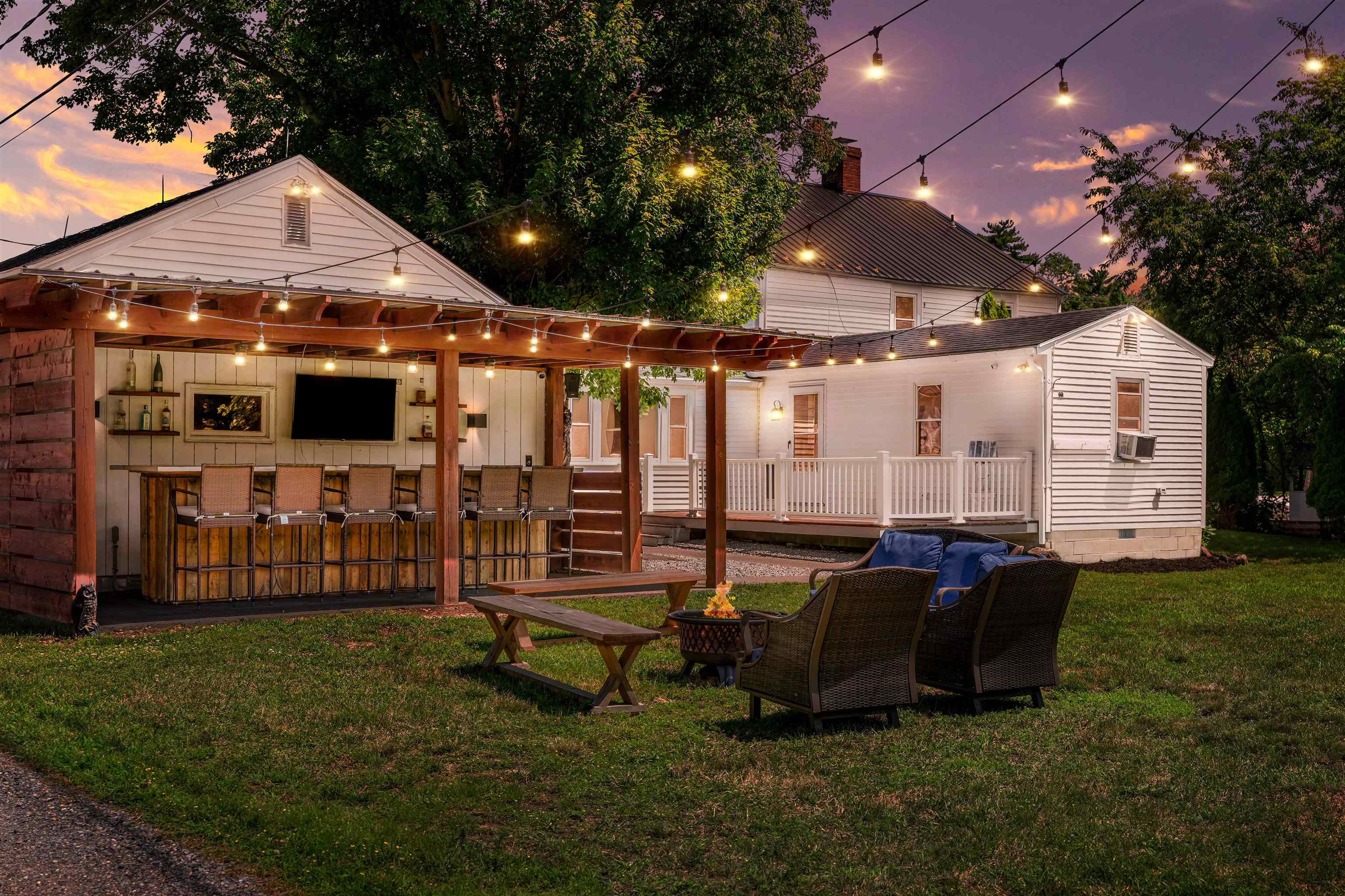 Backyard at dusk with a covered outdoor bar, fire pit, wicker chairs, and string lights illuminating the white house.