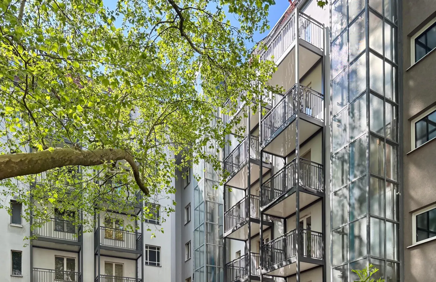 Modern apartment building exterior with stacked metal balconies and a glass elevator shaft, framed by lush green tree leaves.