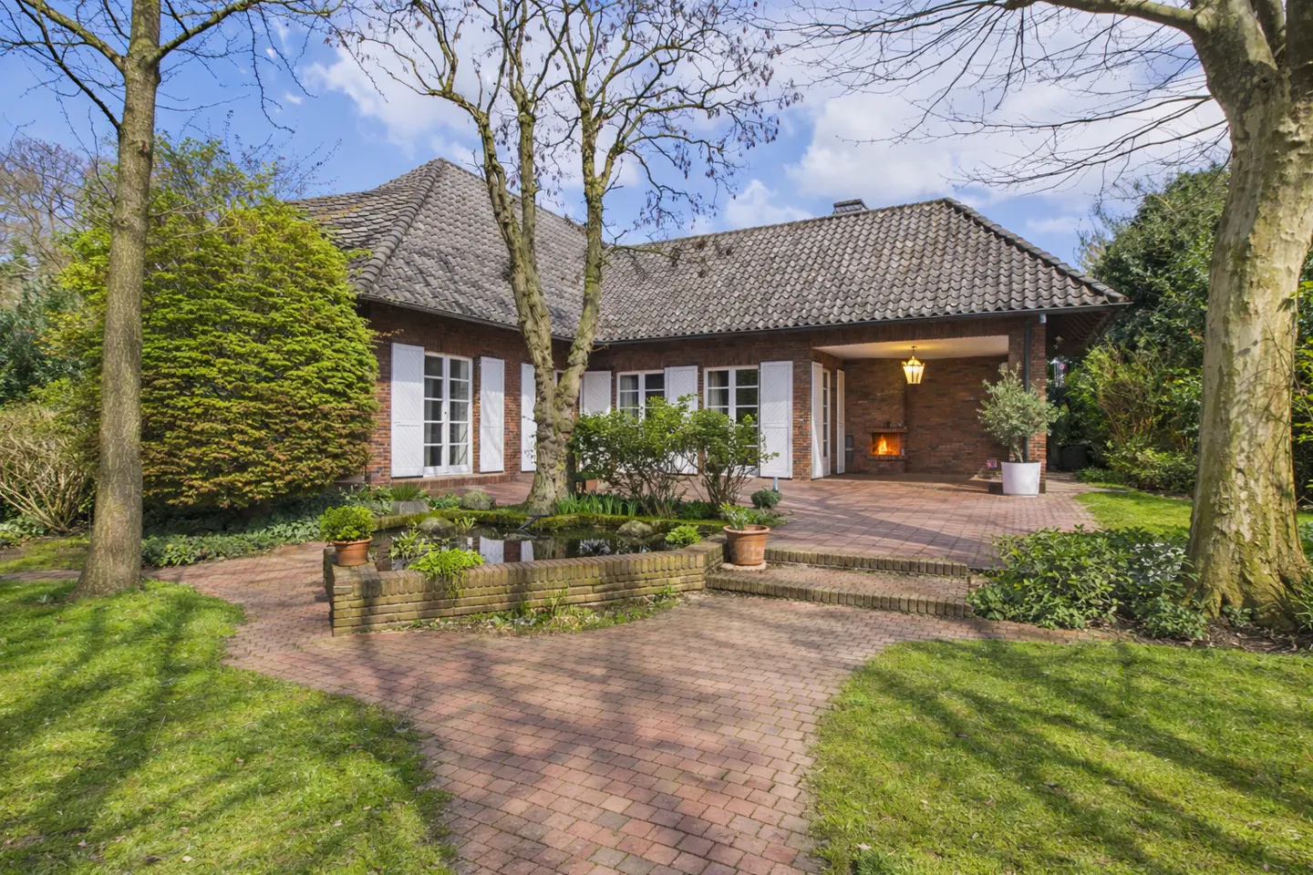 Exterior of a brick house with white shutters, a brick patio, and a pond. A fireplace is visible on the patio.