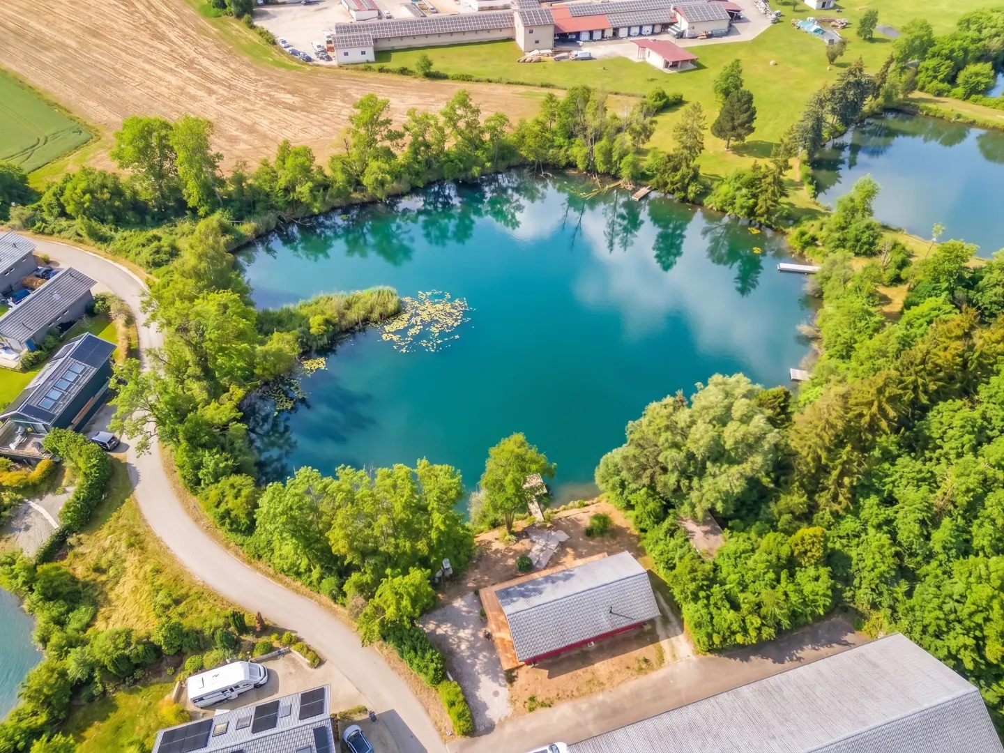 Aerial view of a turquoise lake surrounded by green trees, with buildings and fields in the background.
