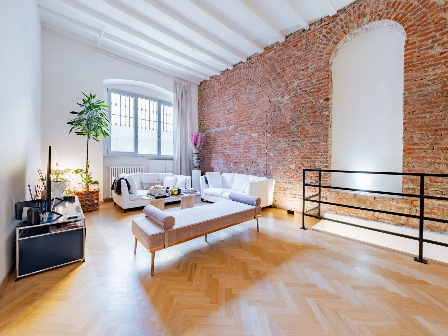 Bright living room with white sofas, pink bench, and exposed brick wall. A large window provides natural light.