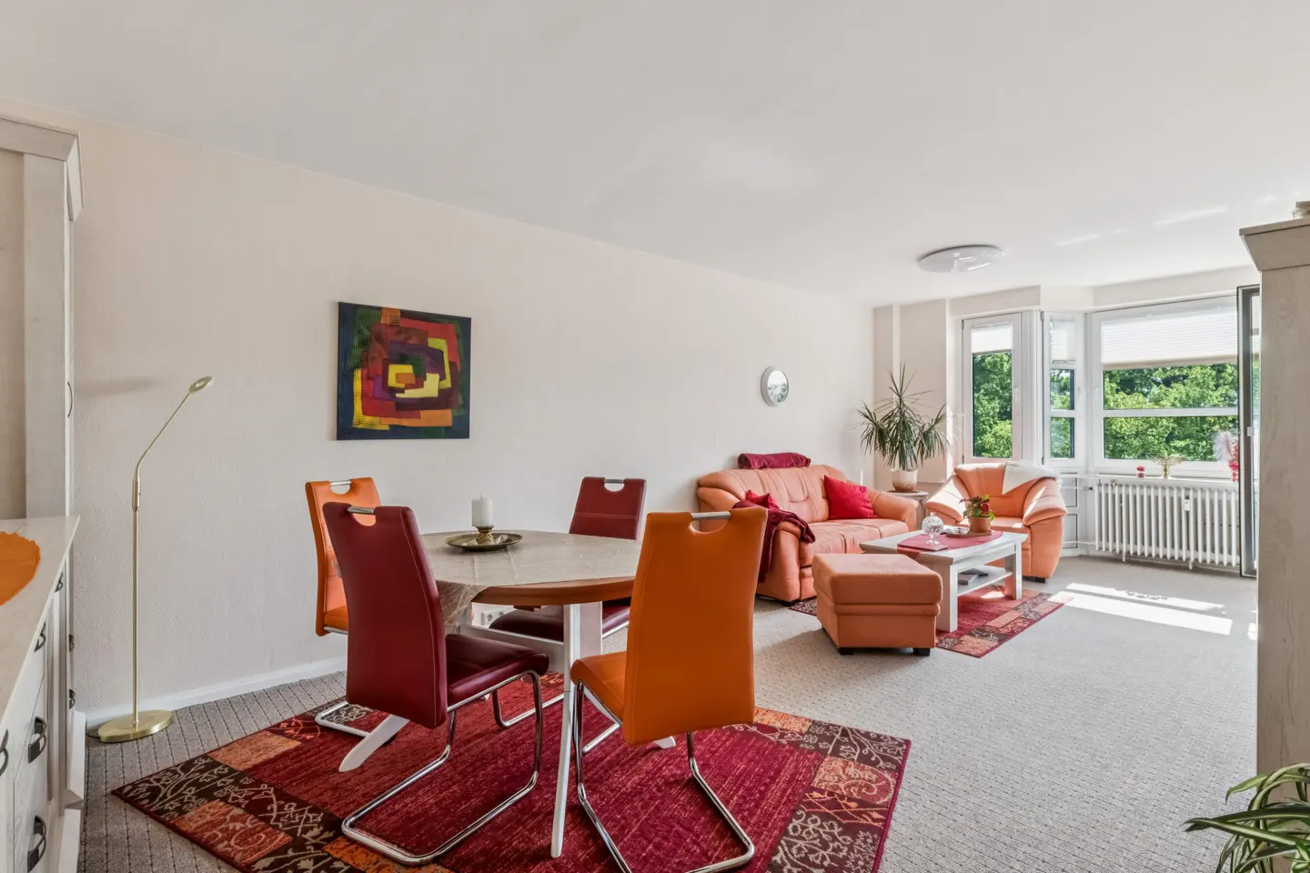 Bright living room with a dining area, orange and red chairs, a matching sofa, and large windows with a garden view.