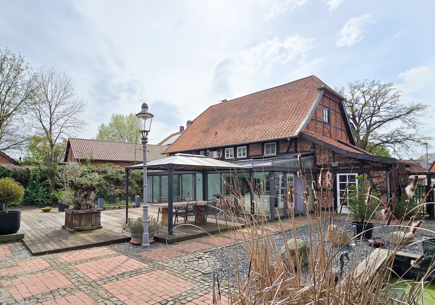 Exterior view of a brick house with a red tile roof, a patio with a gazebo, and a pond with reeds.