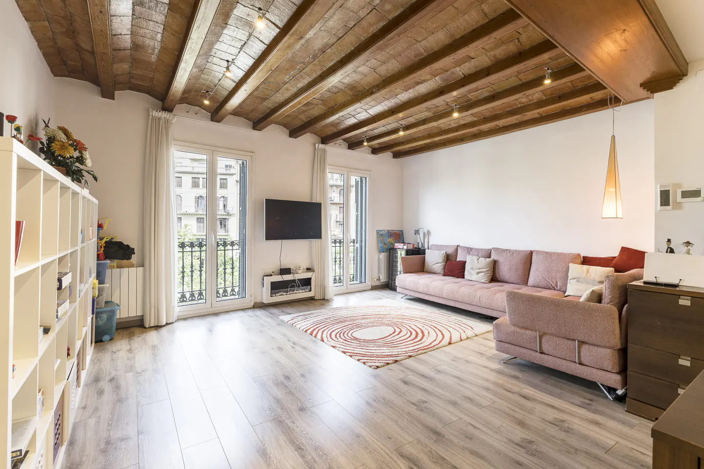 Bright living room with wood floors, exposed beam ceiling, and pink sofa. Balconies with white doors offer city views.