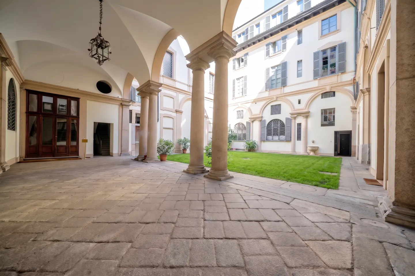 A stone-paved courtyard with classical columns, arched walkways, and a central green lawn surrounded by white buildings.