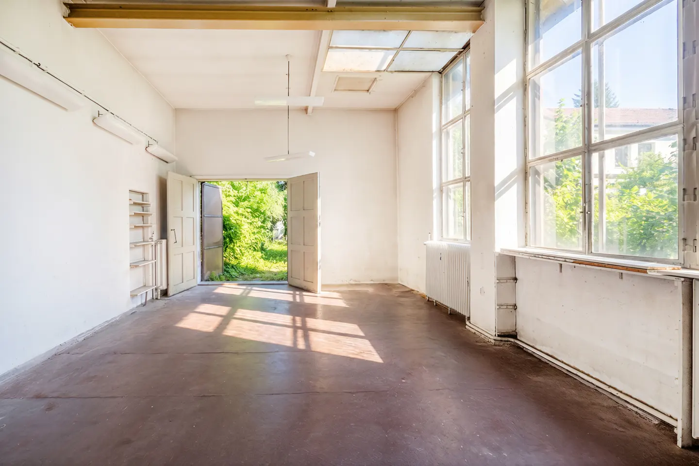 Empty, sunlit industrial room with white walls, brown concrete floors, large windows, and open doors to a green garden.