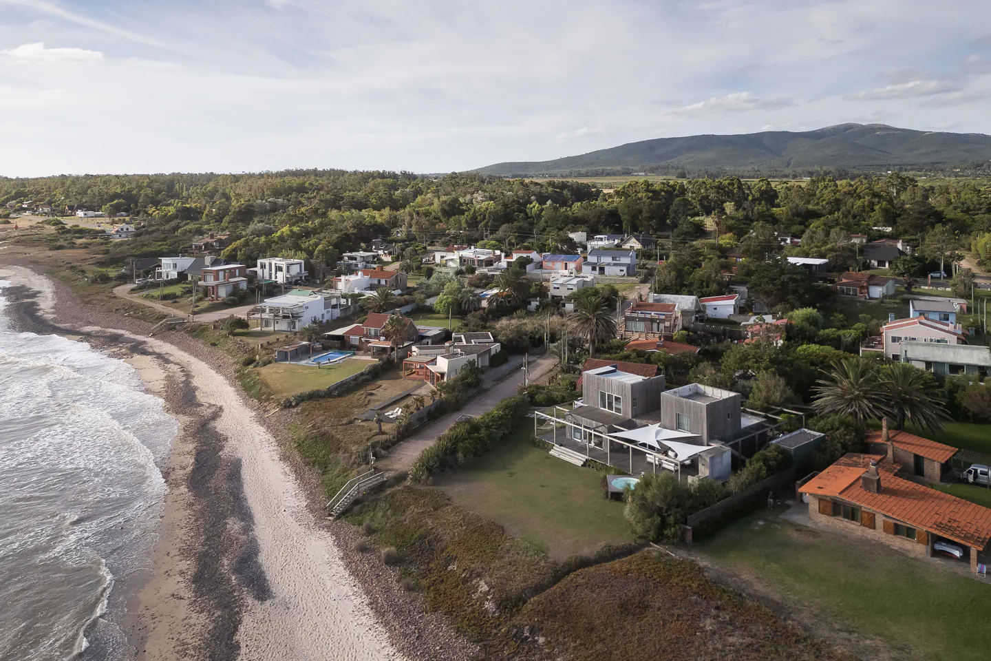 Aerial view of a coastal neighborhood with modern homes, sandy beach, and green trees against a mountain backdrop.