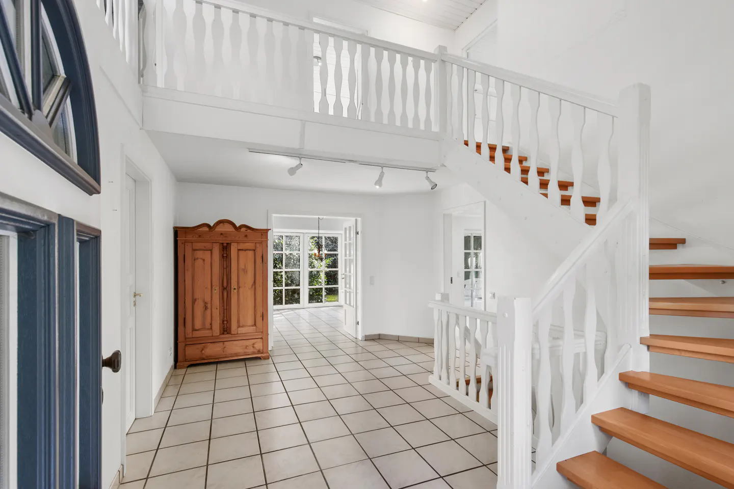Bright foyer with white walls, tile floor, and wooden staircase. A wooden cabinet sits near open doors leading to a garden.