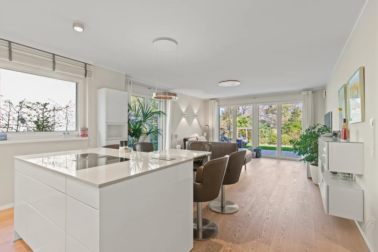 Bright, modern open-concept living space with a white kitchen island, brown bar stools, and wood floors. Large windows offer a view of the green outdoors.