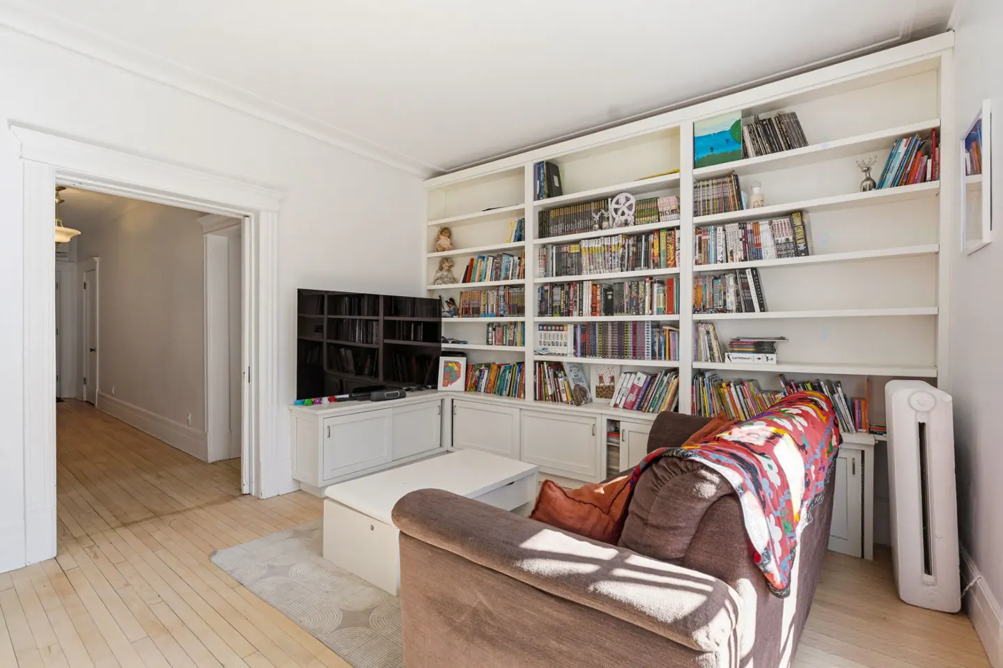 Living room with a brown sofa, white bookshelf filled with books, and a TV on a white cabinet. The room has light wood floors and white walls.