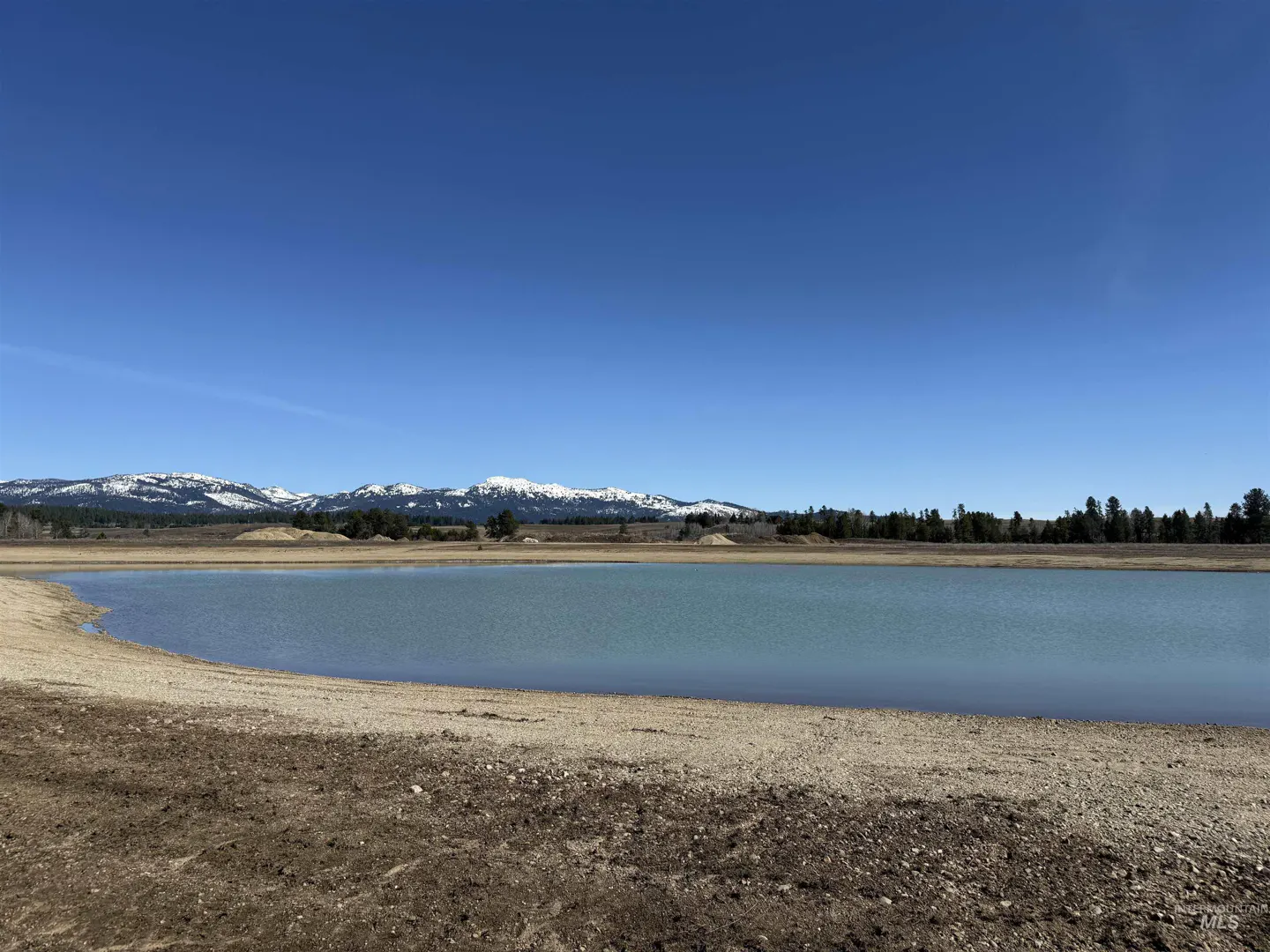A scenic view of a blue lake with snow-capped mountains in the background under a clear blue sky.