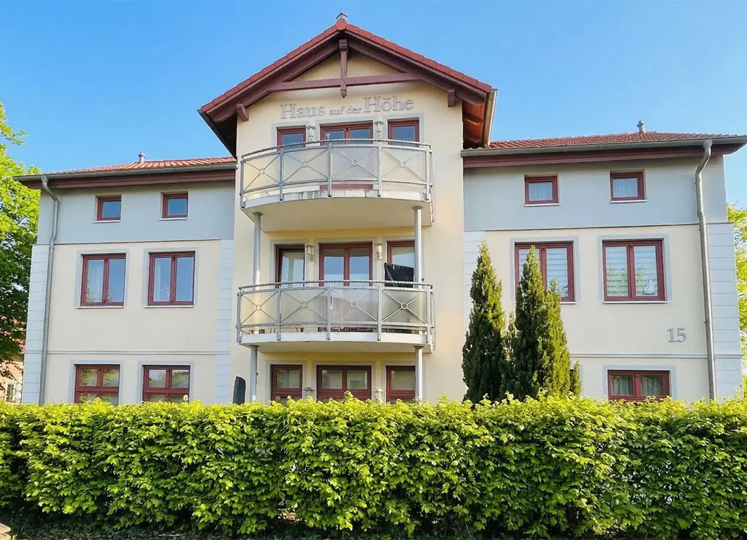 Three-story apartment building with balconies, red-framed windows, and a green hedge in front. "Haus auf der Höhe" is written on the building.