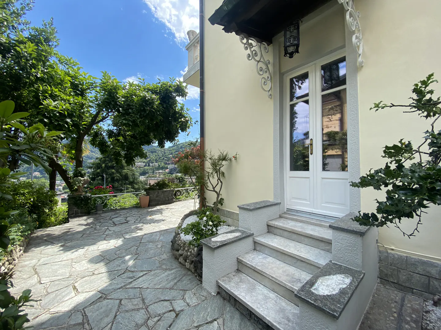 Cream-colored house exterior with white double doors, stone steps, and a paved patio surrounded by lush green trees.