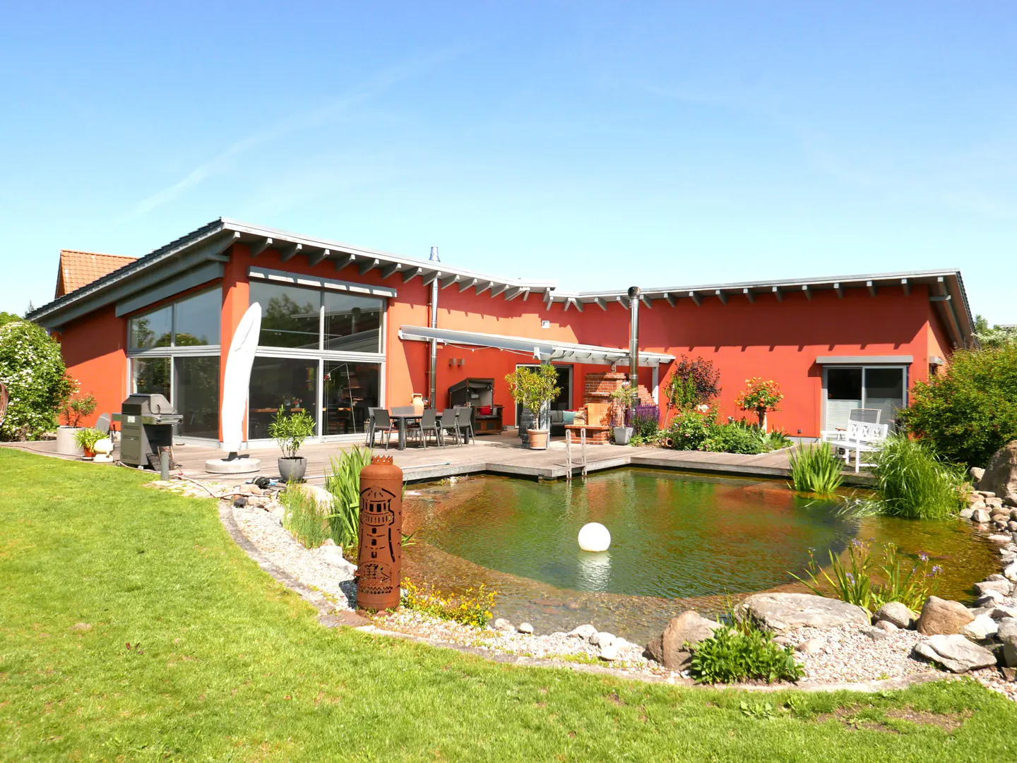 Red house with a pond. The pond is surrounded by a wooden deck, plants, and rocks. There is a table and chairs on the deck.