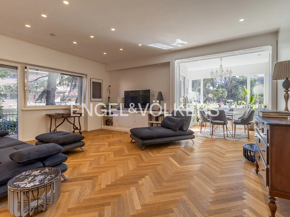 Bright living room with herringbone wood floors, gray sofas, and a dining area with a chandelier. Large windows offer views of greenery.