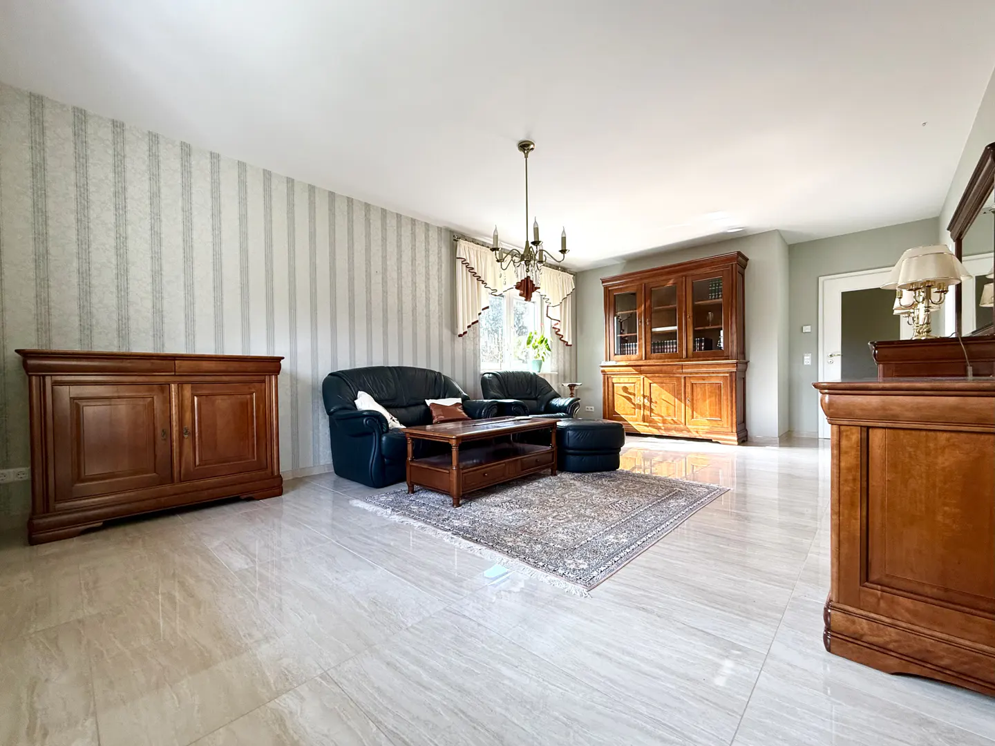 A living room with marble floors, striped wallpaper, and dark wood furniture. Two black leather sofas face a wooden coffee table on a patterned rug. A chandelier hangs from the ceiling.