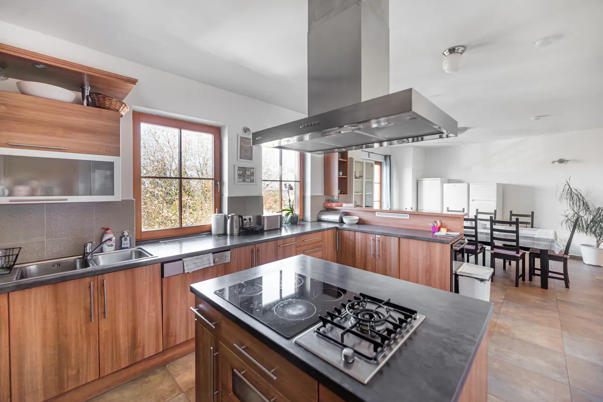 Bright kitchen with wood cabinets, black countertops, and a central island with a gas stovetop. A dining area is visible in the background.