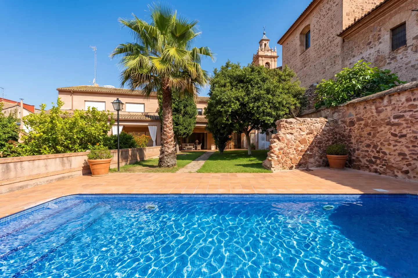 A blue tiled pool with sunlit ripples, a palm tree, and stone buildings under a clear blue sky.