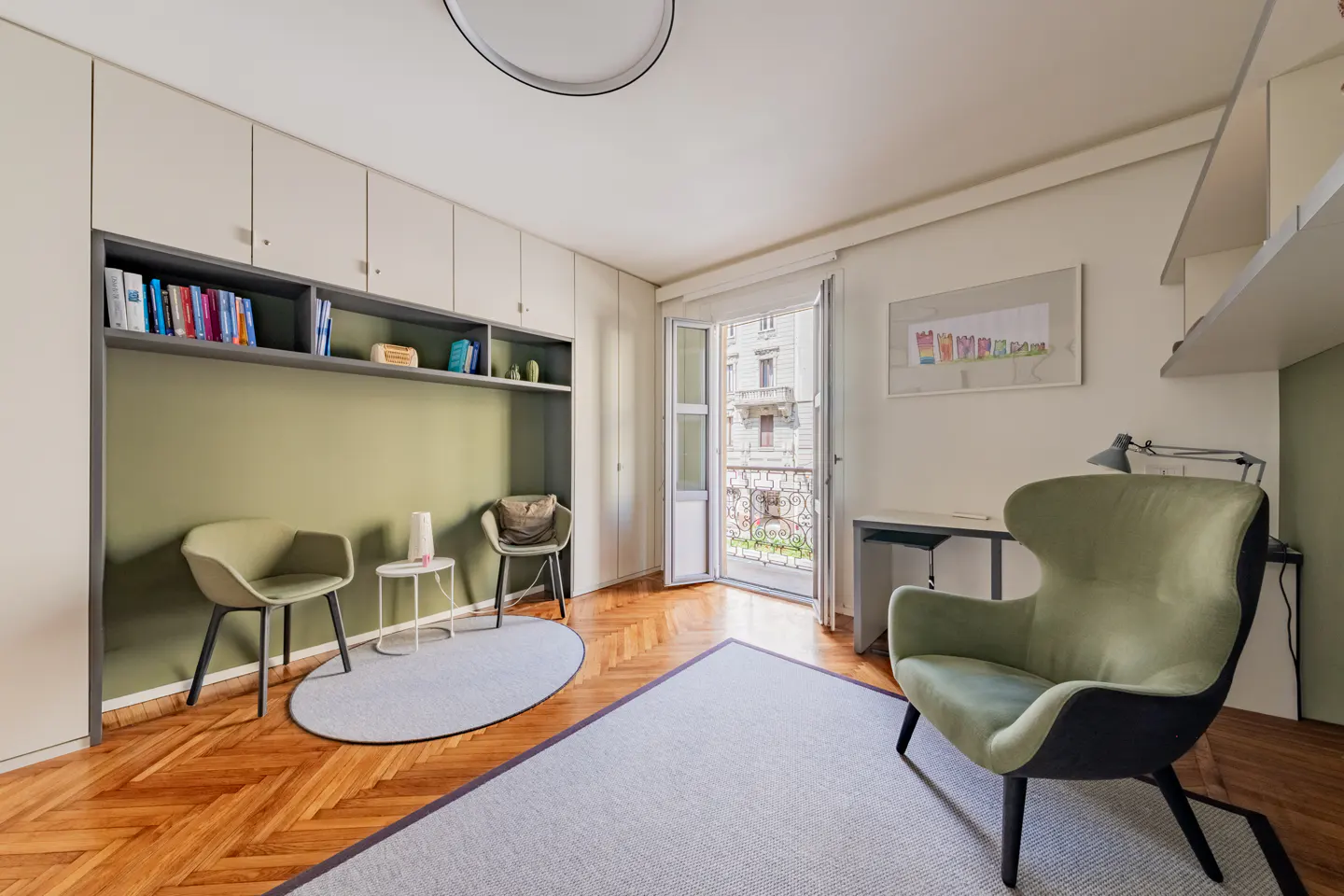 Bright room with hardwood floors, a green accent wall, and built-in shelving. Two chairs and a table sit on a round rug. A desk and open balcony are visible.