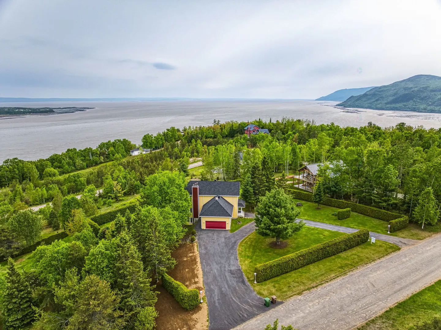 Aerial view of a yellow house with a red garage door, surrounded by green trees and a body of water in the background.