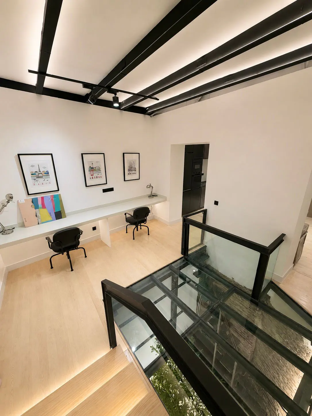 Modern home office with a long white desk, two black chairs, framed art, and a glass floor overlooking the level below.