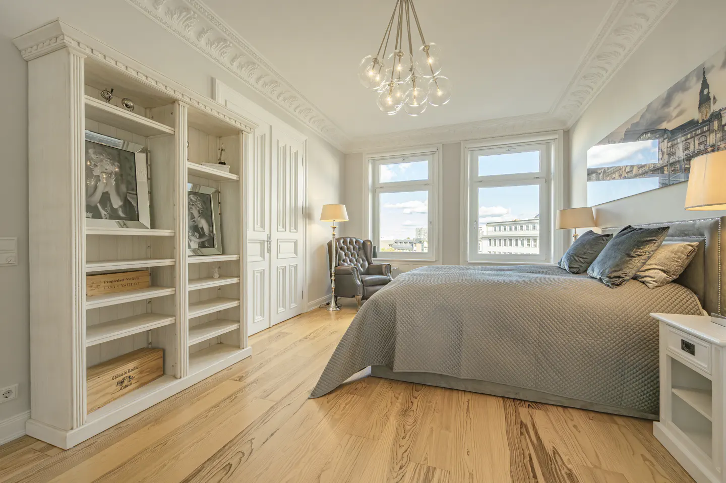 Bright bedroom with wood floors, a grey bed, and a white bookshelf. Windows offer a city view. A modern chandelier hangs from the ceiling.