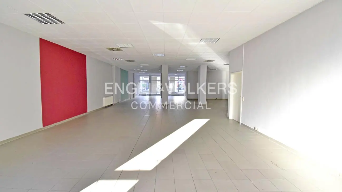 Empty commercial space with white walls, tile floor, and a red accent wall. Columns support the ceiling with recessed lighting.