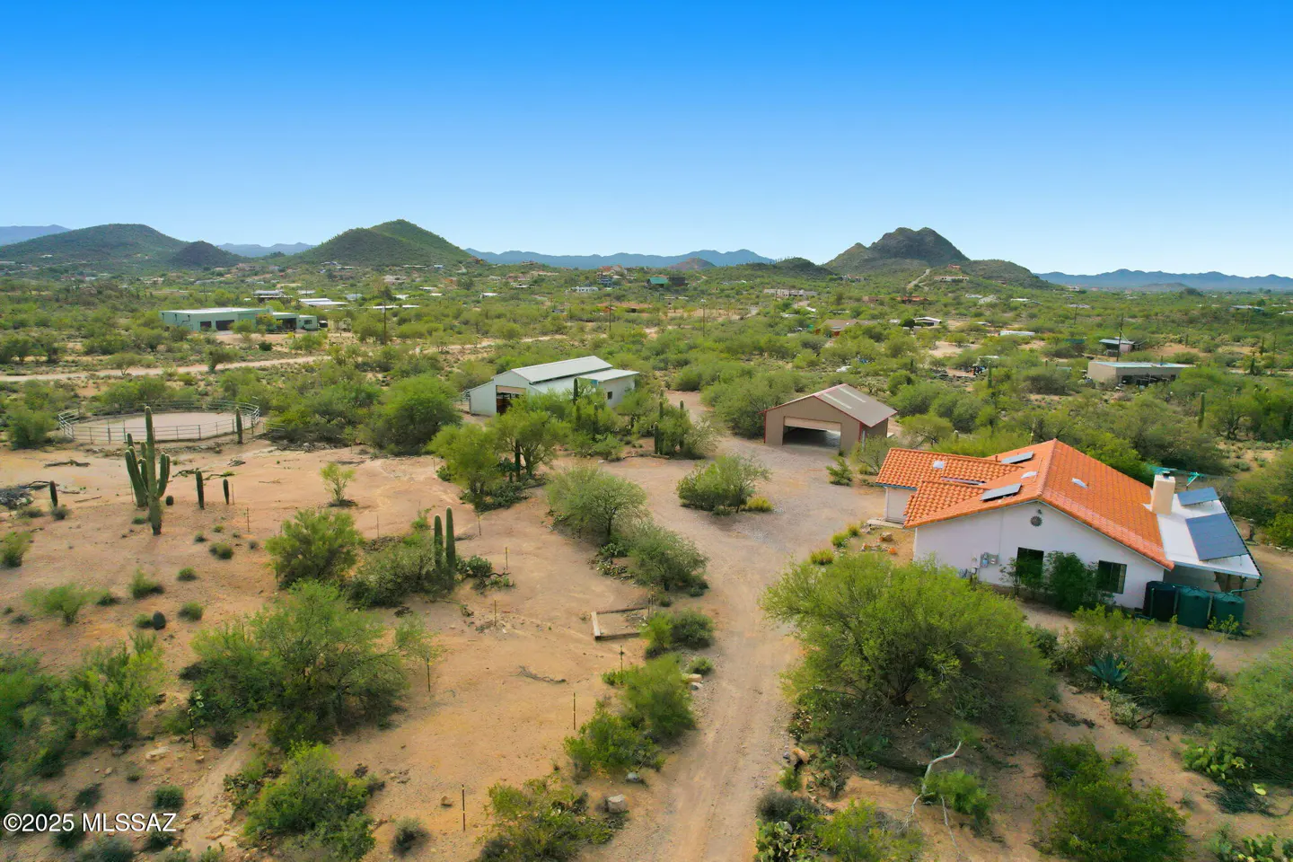 Aerial view of a desert property with a white house, orange roof, solar panels, and outbuildings. Cacti and mountains are in the background.